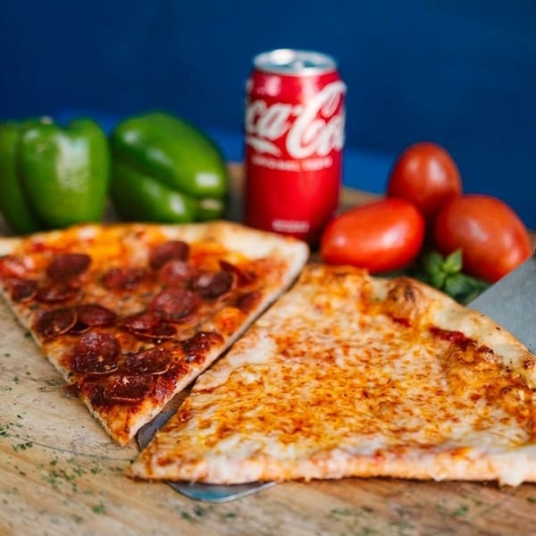 Two pizza slices, one pepperoni and one cheese, with a soda, peppers, and tomatoes on a board.