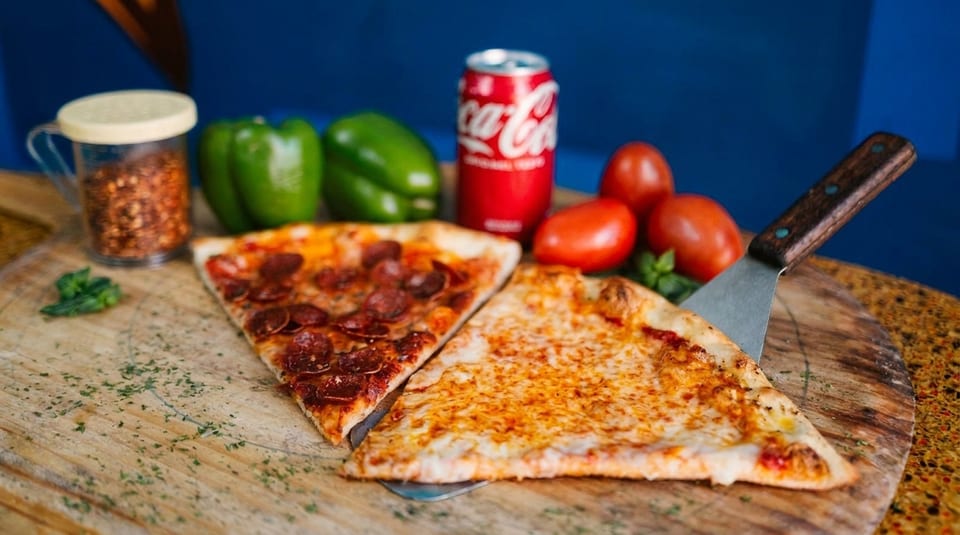Two pizza slices, one pepperoni and one cheese, with a soda, peppers, and tomatoes on a board.