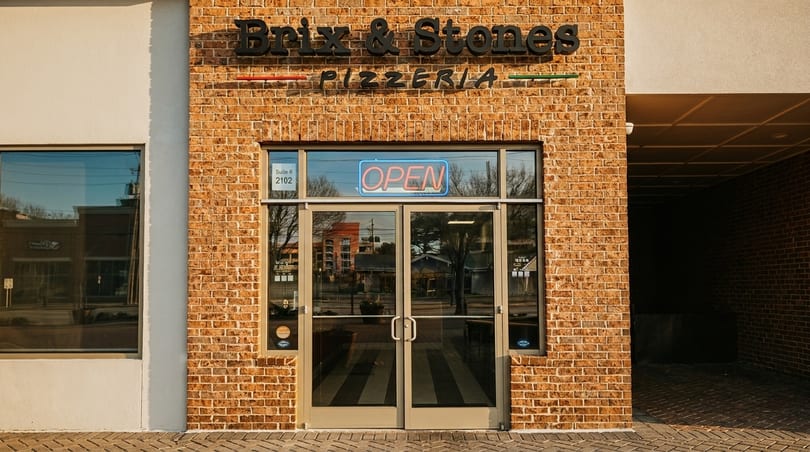 Exterior of Brix & Stones Pizzeria with a brick facade, glass doors, and an "OPEN" neon sign.