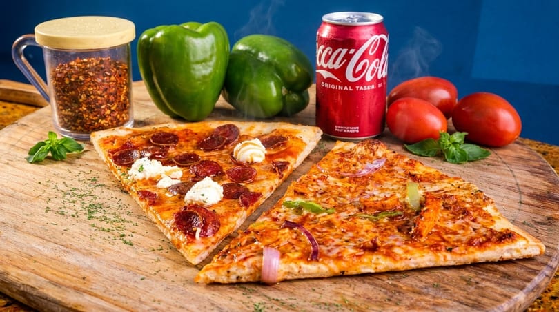 Two pizza slices, a Coca-Cola can, green peppers, tomatoes, and chili flakes on a wooden board.