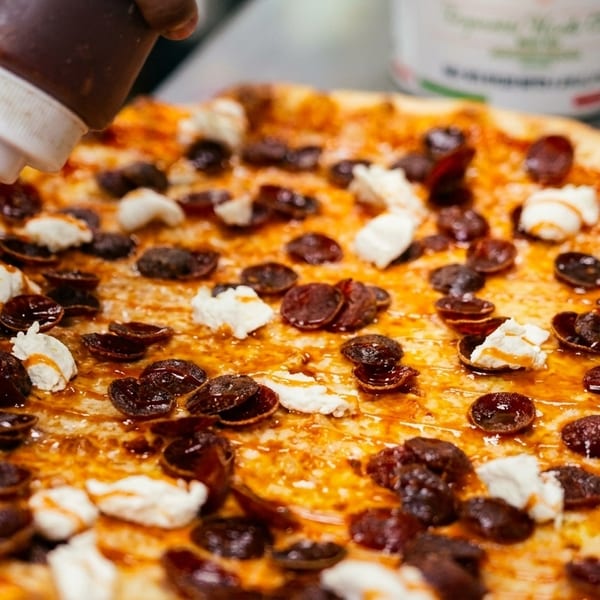 A chef drizzles sauce onto a pizza topped with pepperoni and dollops of cheese.