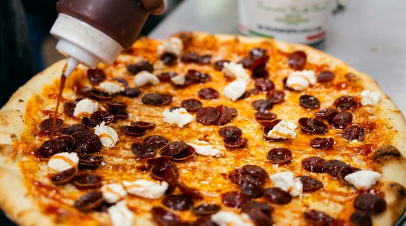 A chef drizzles sauce onto a pizza topped with pepperoni and dollops of ricotta cheese.