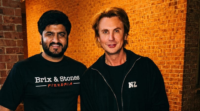 Two men, one in a Brix & Stones Pizzeria shirt, smiling against a brick wall.