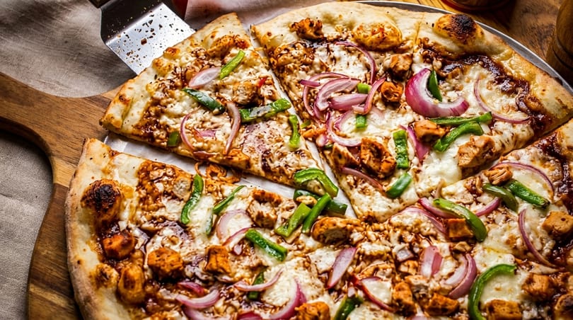 A BBQ chicken pizza with red onions and green peppers, with a slice being served.